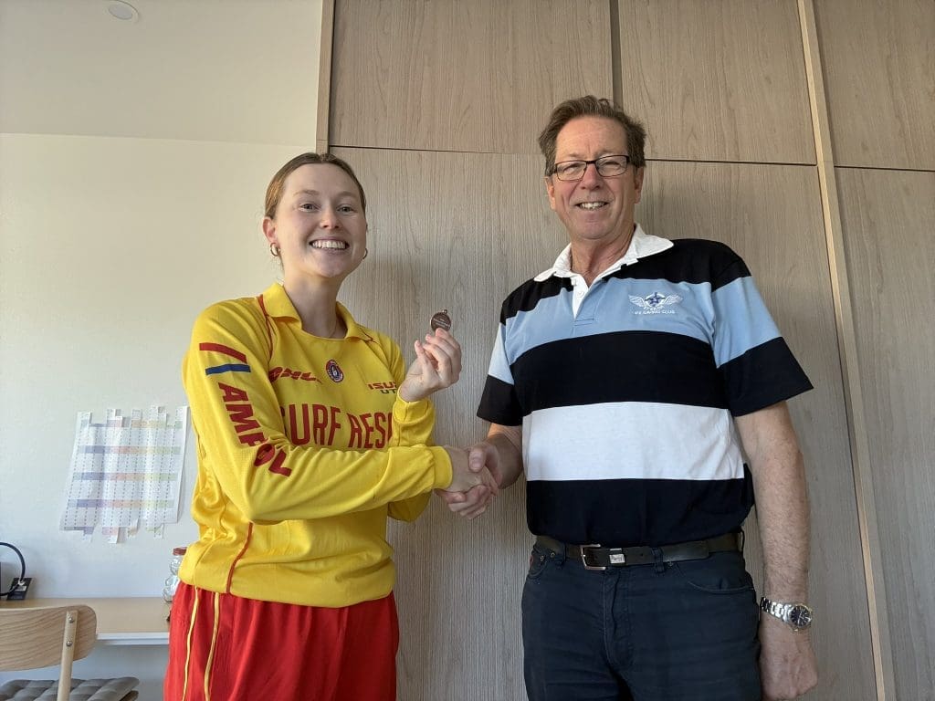 A young surf lifesaver in a yellow Surf Rescue uniform shakes hands with a smiling male club member indoors while holding a medal